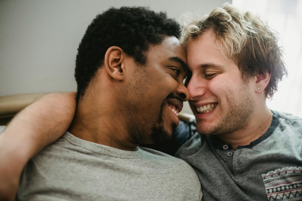 A happy interracial same-sex couple embracing each other with smiles indoors.